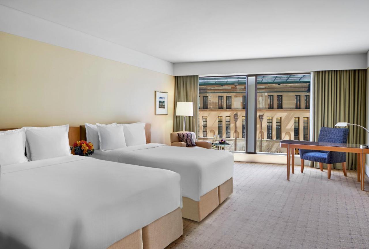 더 풀러턴 호텔 시드니 외부 사진 The photo shows a modern hotel room with two neatly made beds, each adorned with white bedding and pillows. There is a bedside table with a small decorative arrangement and a lamp beside one of the beds. The room features large windows with sheer cur