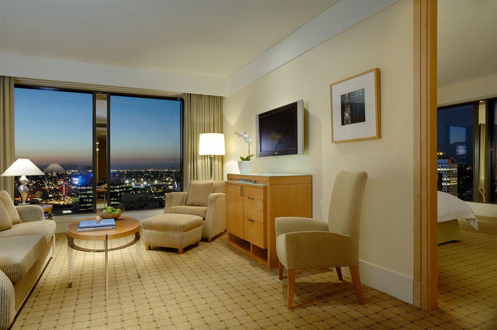 더 풀러턴 호텔 시드니 외부 사진 The photo shows a hotel room with a modern and elegant design. There's a large window with a view of a city skyline, indicating it's likely at a higher floor. The room features a seating area with two light-colored chairs and a small round coffee tab