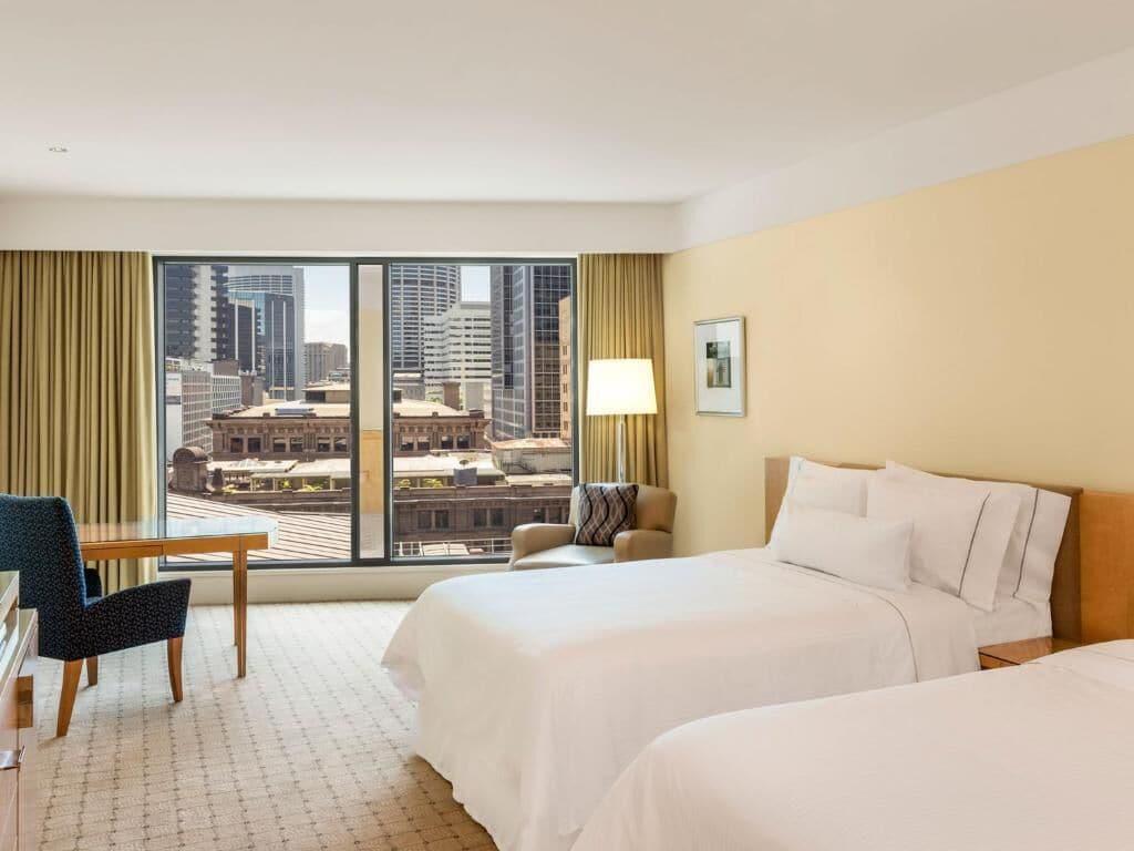 더 풀러턴 호텔 시드니 외부 사진 The photo shows a hotel room with a modern design. There are two beds with white linens positioned in the center. A large window offers a view of a cityscape, featuring tall buildings outside. To the side, there is a comfortable armchair and a wooden