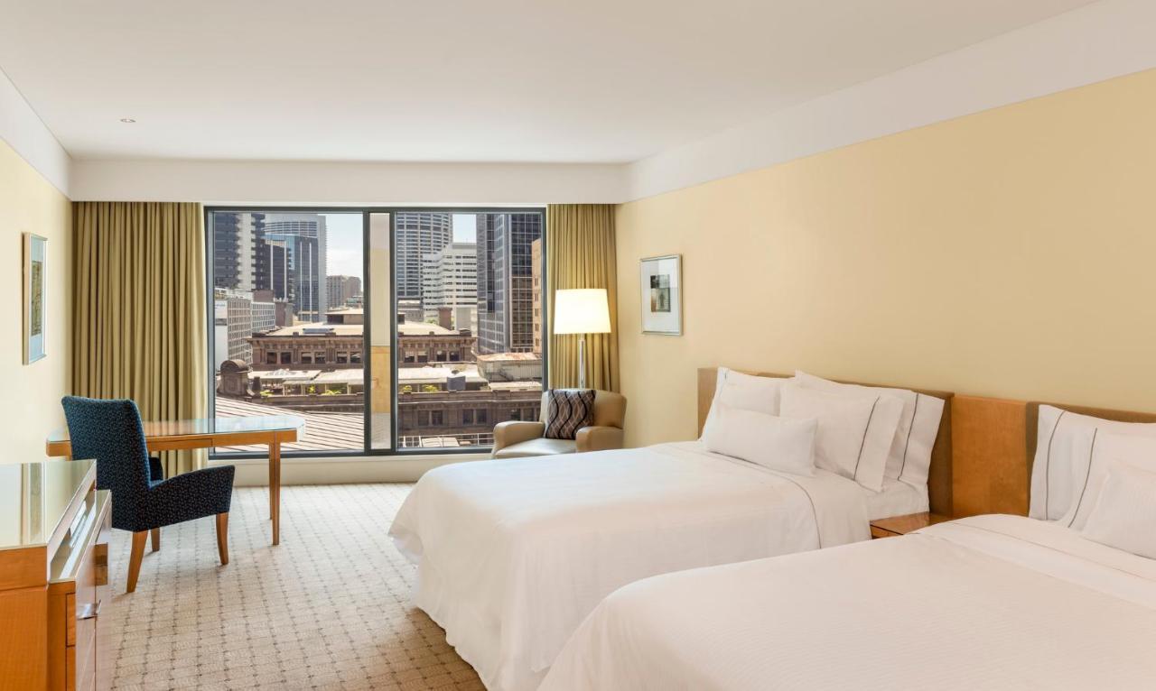 더 풀러턴 호텔 시드니 외부 사진 The photo shows a hotel room featuring two beds with white bedding. The walls are painted a soft yellow, creating a warm atmosphere. Large windows provide a view of the city skyline, and there are beige curtains framing the windows. In the room, ther