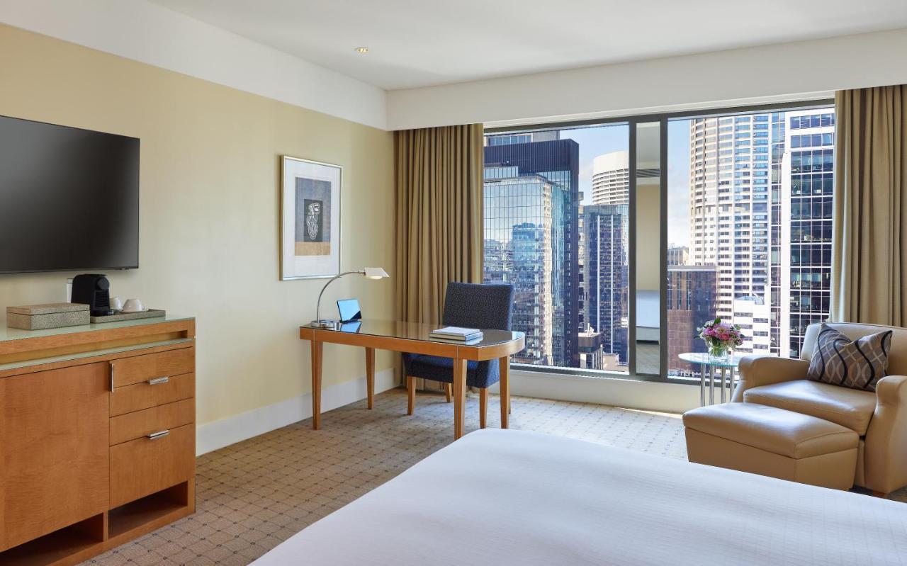 더 풀러턴 호텔 시드니 외부 사진 The photo shows a modern hotel room with a cozy and contemporary design. There is a comfortable bed with white linens, a desk with a chair and a lamp positioned by a large window that offers a view of a cityscape filled with tall buildings. A flat-sc
