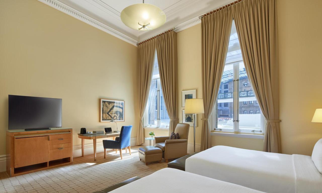 더 풀러턴 호텔 시드니 외부 사진 The photo shows a hotel room featuring a light-colored interior with high ceilings. There are two beds positioned at the forefront of the image, with a small bedside table between them. To one side, a wooden dresser supports a flat-screen television.