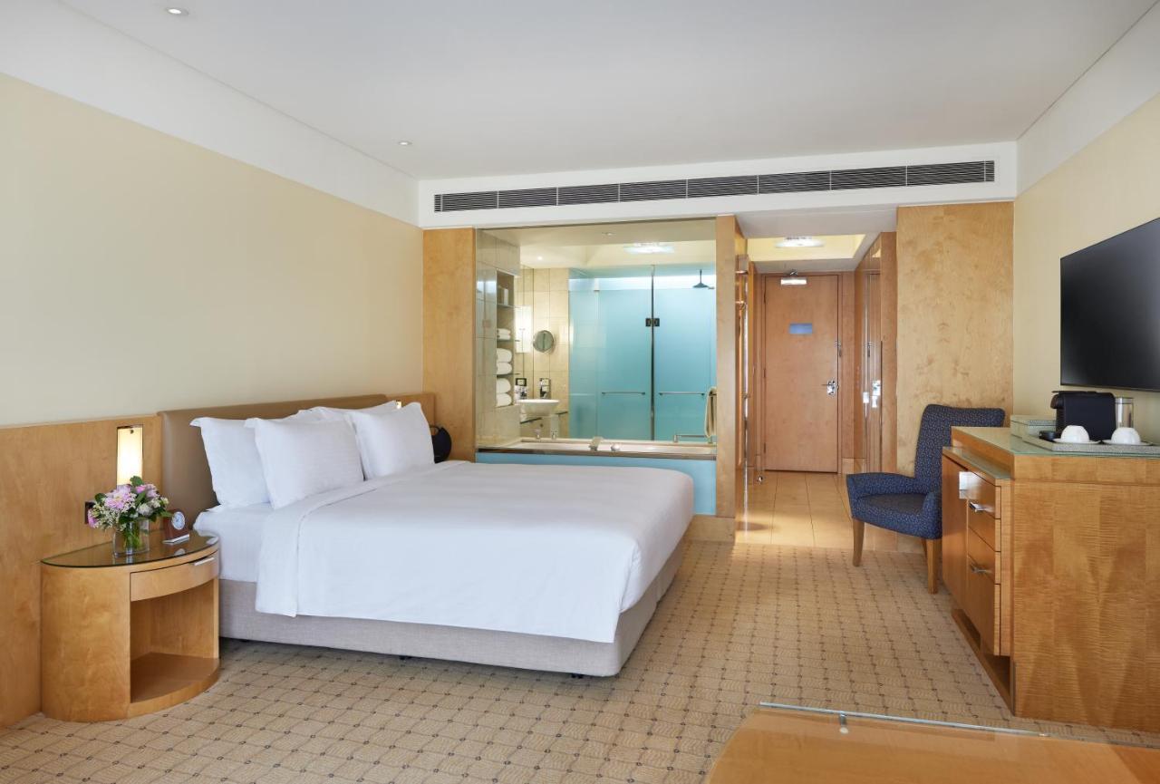 더 풀러턴 호텔 시드니 외부 사진 The photo shows a modern hotel room featuring a spacious layout. It includes a large bed with white linens, a wooden bedside table with a flower arrangement, and a chair in the corner. The room has a light-colored, neutral palette, giving it a clean 
