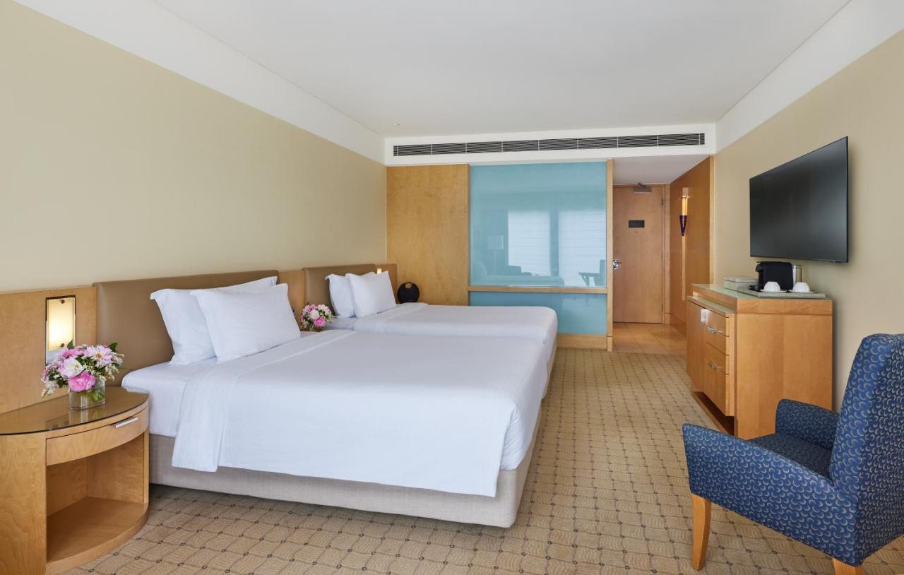 더 풀러턴 호텔 시드니 외부 사진 The image shows a hotel room featuring two twin beds with white linens and decorative pillows. The walls are painted in a soft beige color, and there is wooden furniture, including a bedside table and a media console. A flat-screen TV is mounted on t