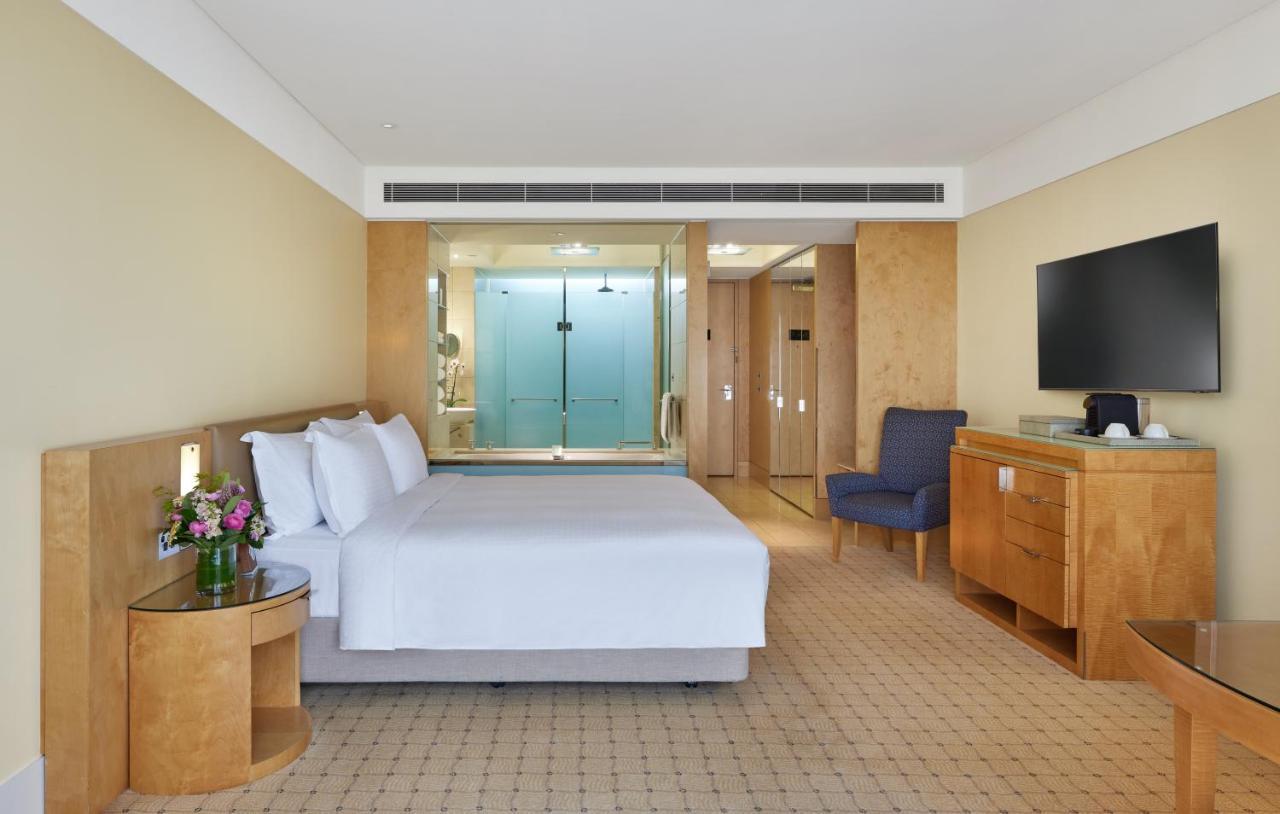 더 풀러턴 호텔 시드니 외부 사진 The photo shows a modern hotel room. It features a large bed with white linens, a small bedside table with a vase of flowers, and a comfortable blue armchair in one corner. There is a wooden dresser and a flat-screen TV mounted on the wall. Behind th