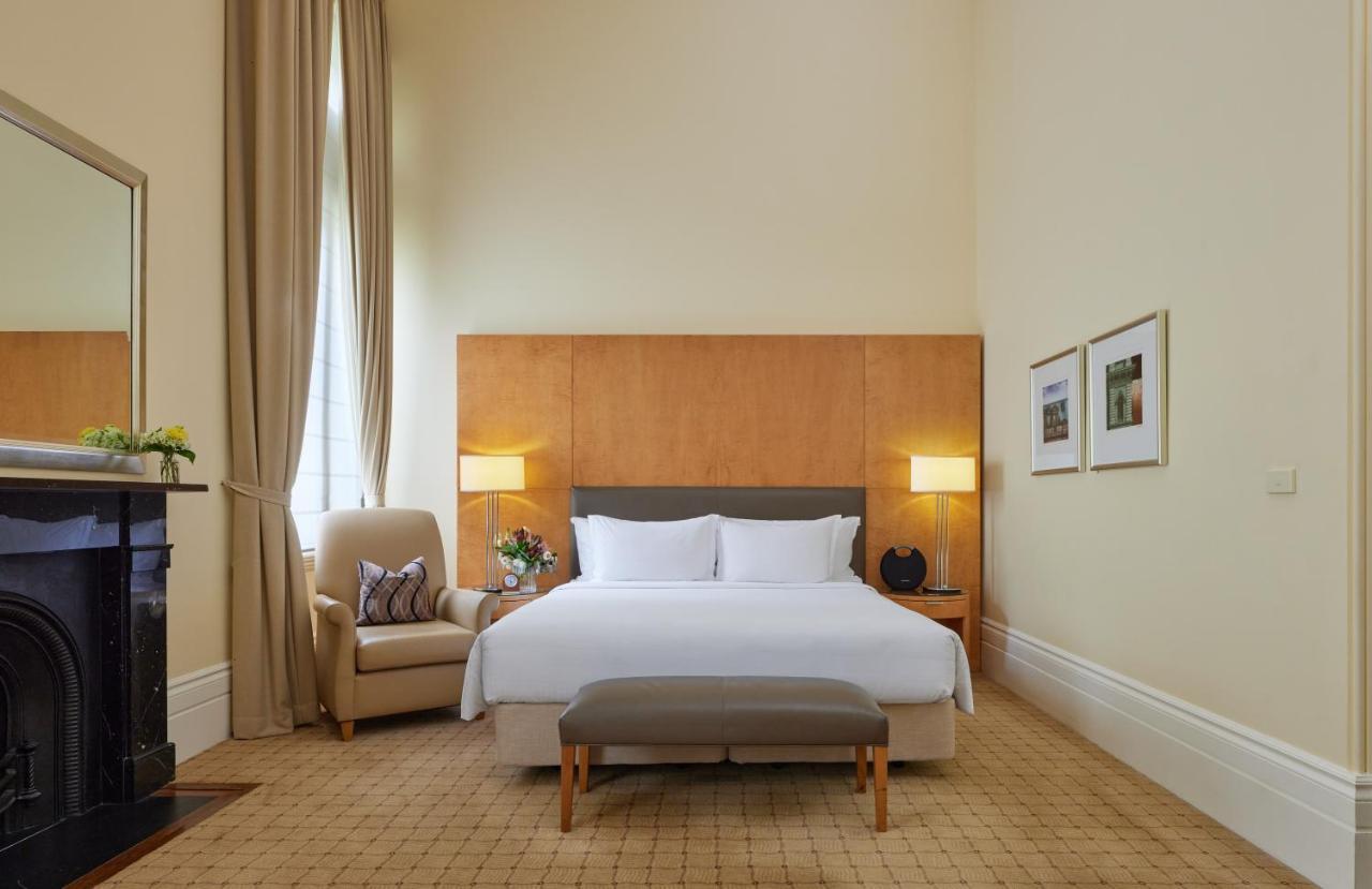 더 풀러턴 호텔 시드니 외부 사진 The photo shows a well-appointed hotel room. It features a large bed with white linens and a gray headboard, positioned against a wooden wall panel. On either side of the bed are stylish lamps. There is a comfortable armchair in the corner, accompani