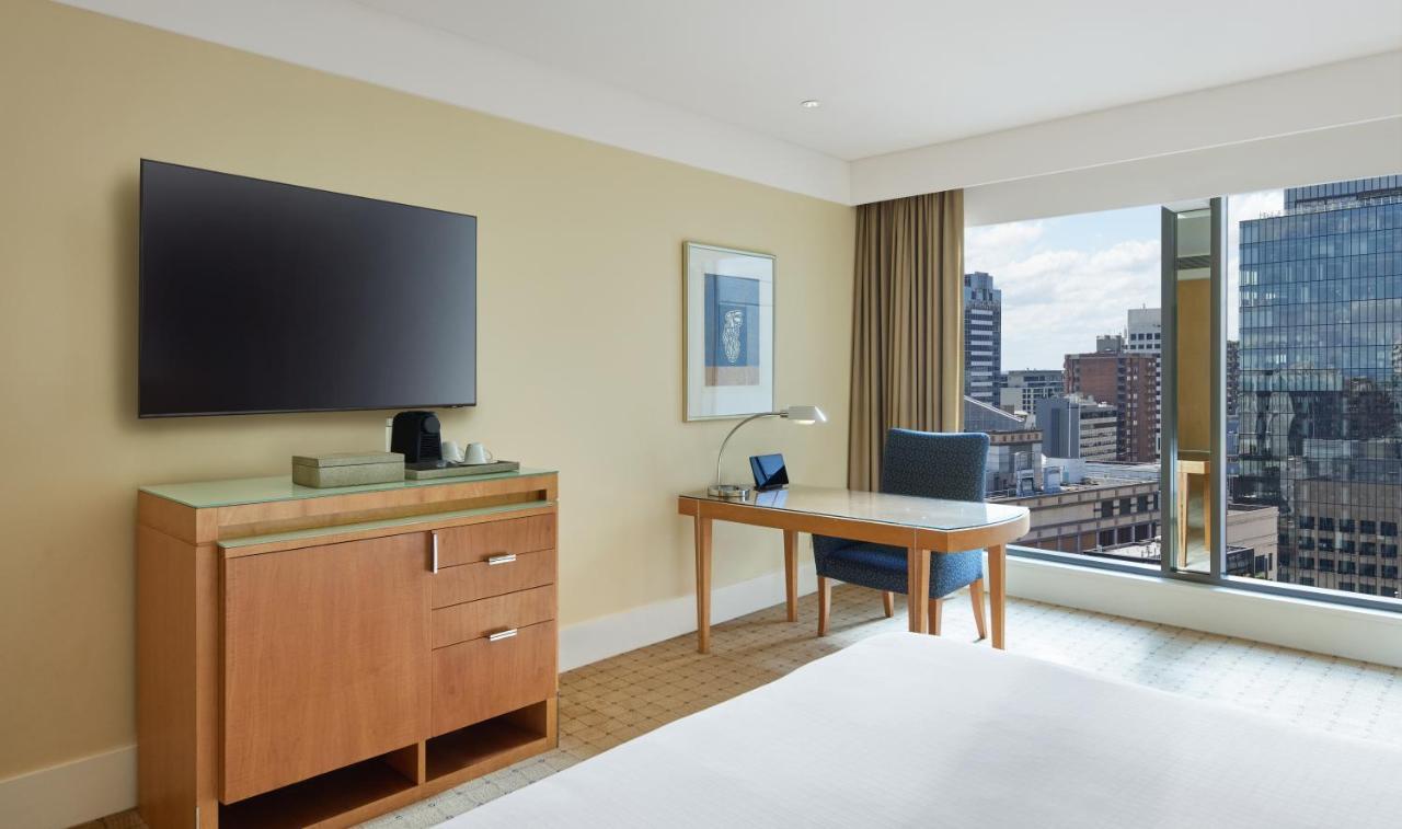 더 풀러턴 호텔 시드니 외부 사진 The photo displays a modern hotel room with a clean and contemporary design. It features a large flat-screen television mounted on the wall. There is a wooden dresser with drawers beneath the TV. A small desk with a lamp and a chair is positioned nea