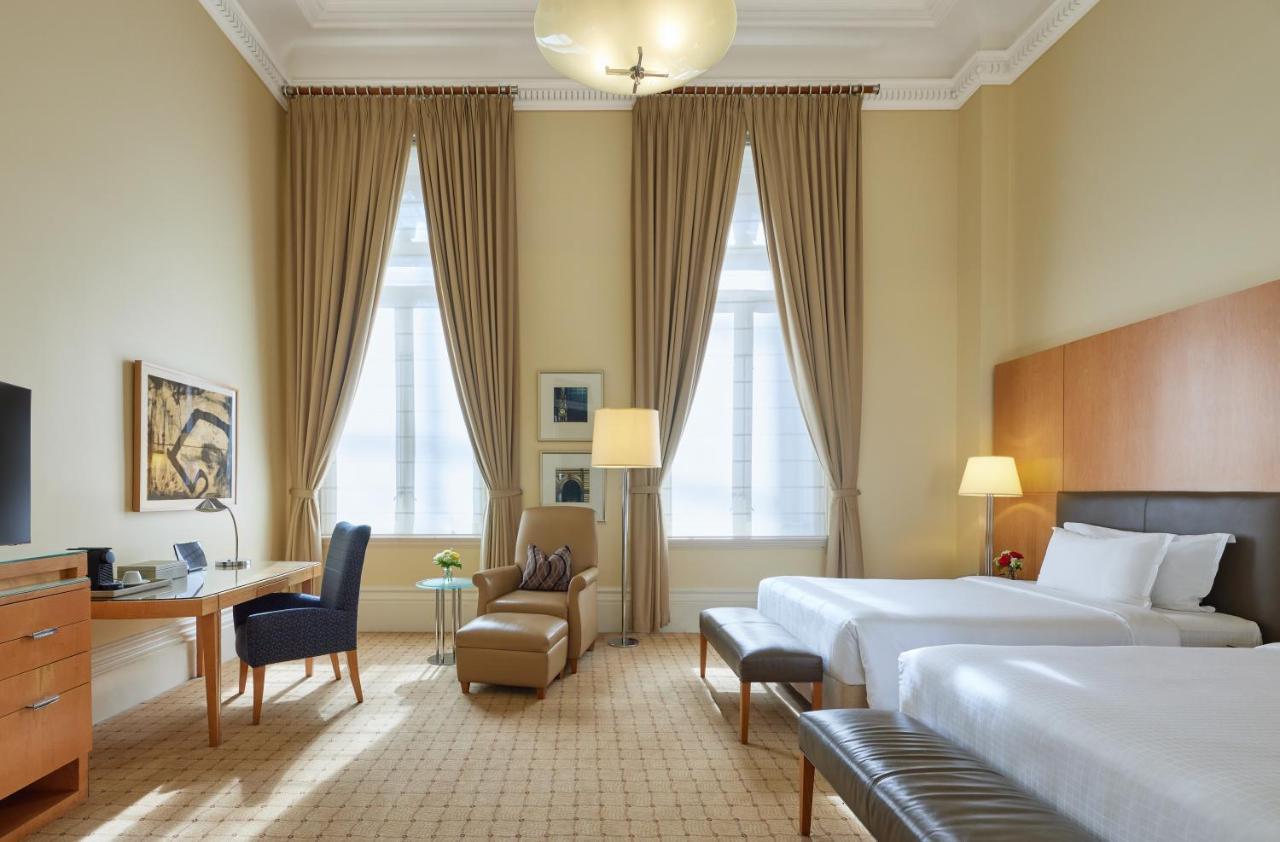 더 풀러턴 호텔 시드니 외부 사진 The photo shows a well-appointed hotel room featuring two beds. The room has high ceilings and large windows dressed with elegant drapes that allow natural light to fill the space. There is a stylish desk and chair positioned against one wall, alongs