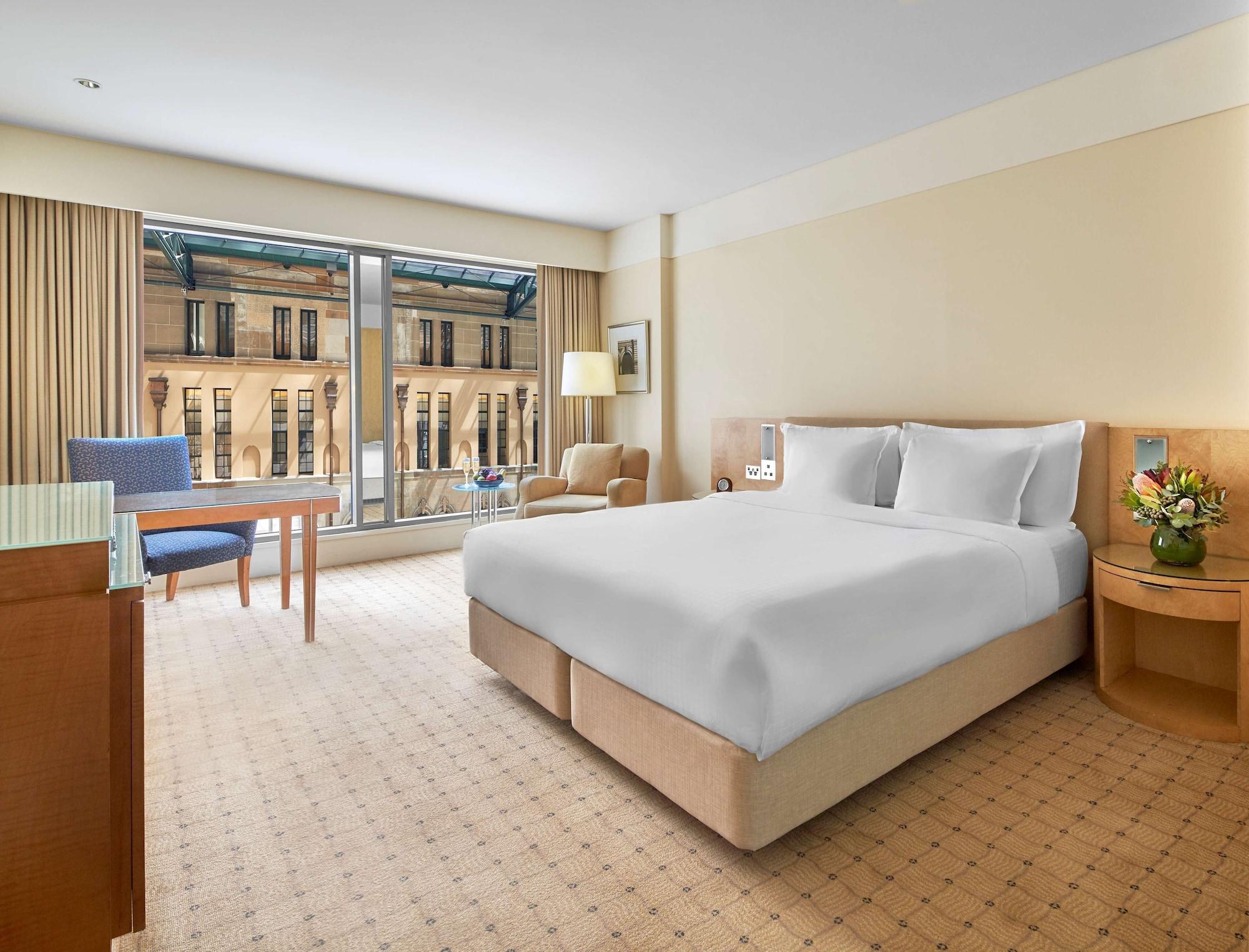 더 풀러턴 호텔 시드니 외부 사진 The photo shows a hotel room featuring a large bed with white bedding at the center. On one side of the room, there is a light-colored bedside table with a lamp. Across from the bed is a wooden table accompanied by a blue chair, while a comfortable a