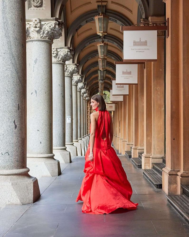 더 풀러턴 호텔 시드니 외부 사진 The photo depicts a woman wearing an elegant red gown, standing in a covered archway lined with columns. She is turned slightly to look back over her shoulder, showcasing the flow and design of the dress. The environment features multiple arches and 