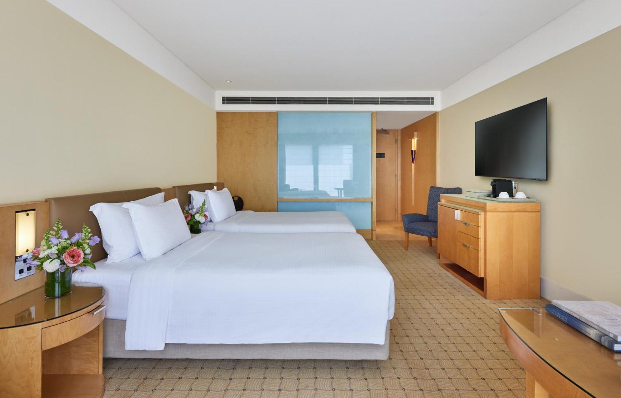 더 풀러턴 호텔 시드니 외부 사진 The photo shows a hotel room with two beds, each made up with white linens and pillows. There are small flower arrangements on the side tables next to the beds. The room has a light color scheme with beige walls and wooden furniture. A blue glass par