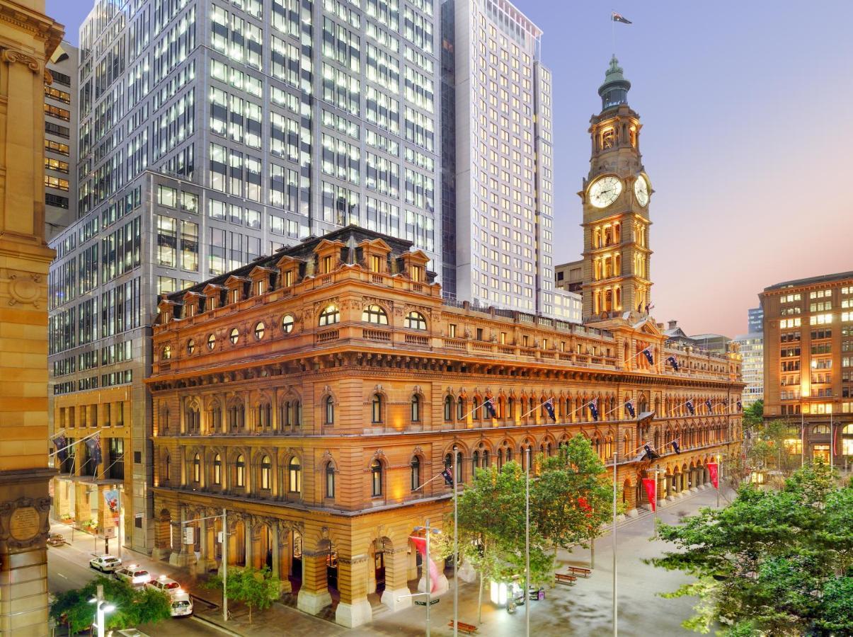 더 풀러턴 호텔 시드니 외부 사진 The photo depicts a cityscape featuring a historical building with ornate architecture, prominently displaying a large clock tower. This structure is set against the backdrop of modern skyscrapers, highlighting a contrast between classic and contempo